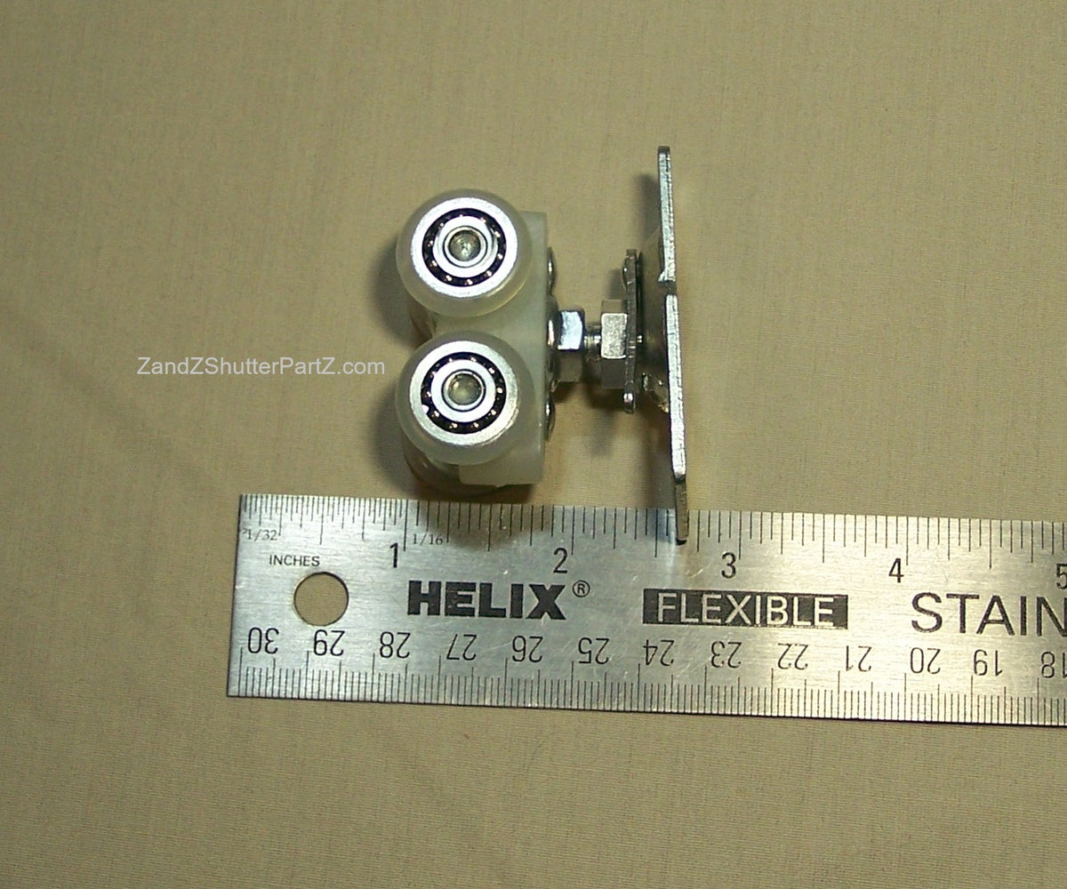 A metal roller assembly part with two wheel bearings and a mounting plate, shown next to a ruler on a beige background.